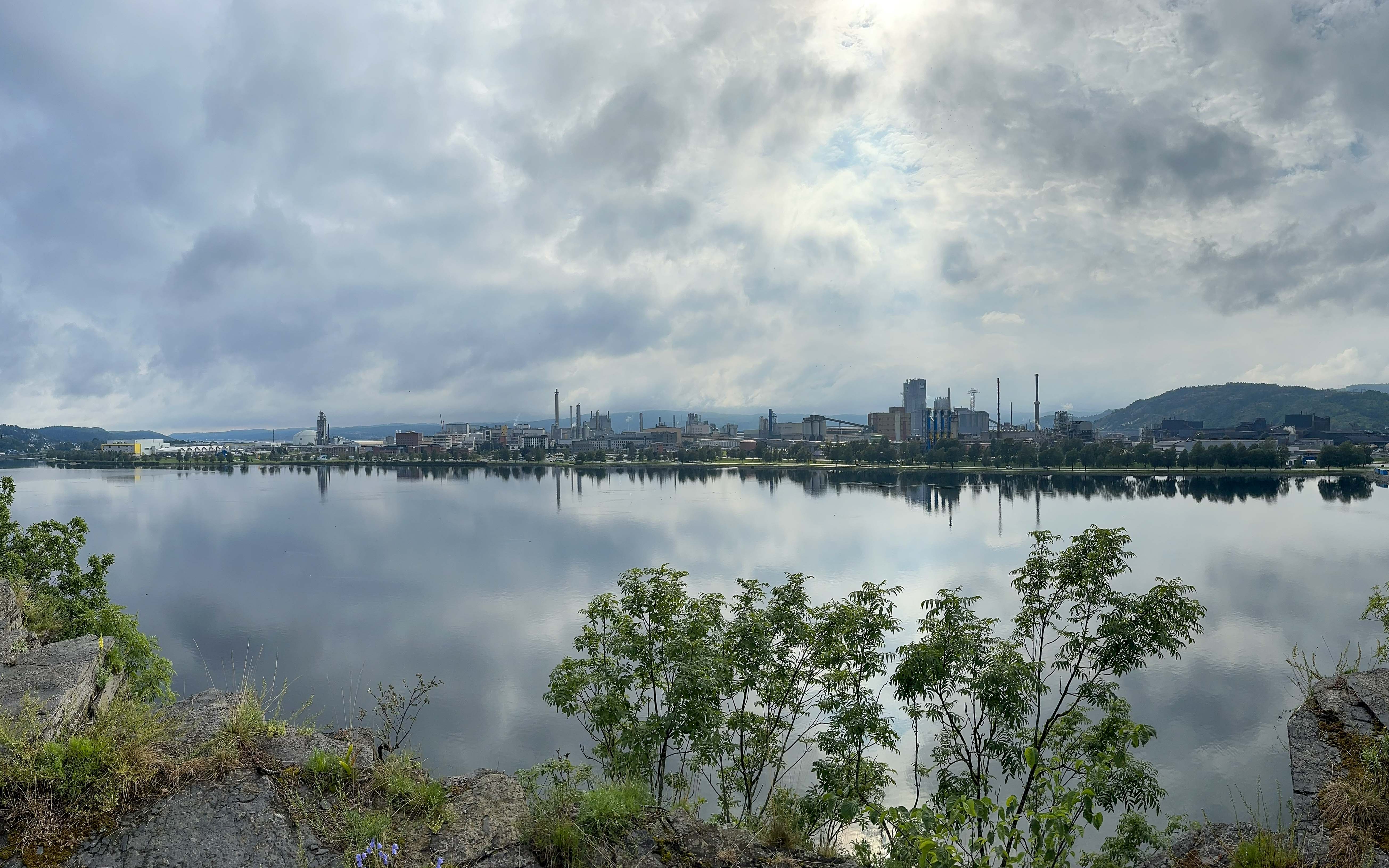 sjø med trær og et industriområde i bakgrunnen
